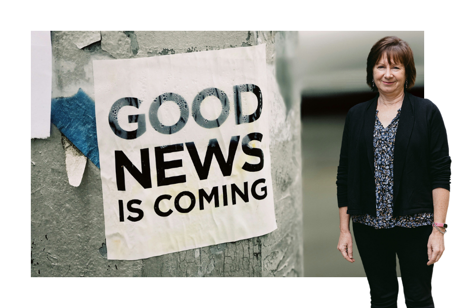 Pictured: Nancy Robertson standing smiling in a black cardigan with a floral shirt underneath and black trousers. Behind her is an image of a sign taped to a power pole that reads; "Good News Is Coming".