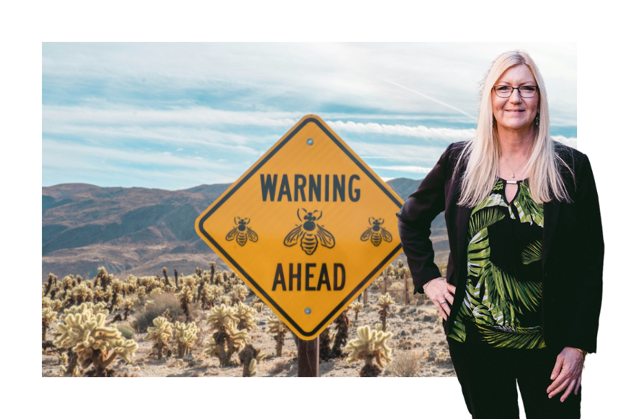 Pictured, Julie Pullen wearing a black blazer and trousers with a beautiful green coloured top with one hand on her hip. Behind her is an image of a yellow road sign that reads "warning, bees ahead".