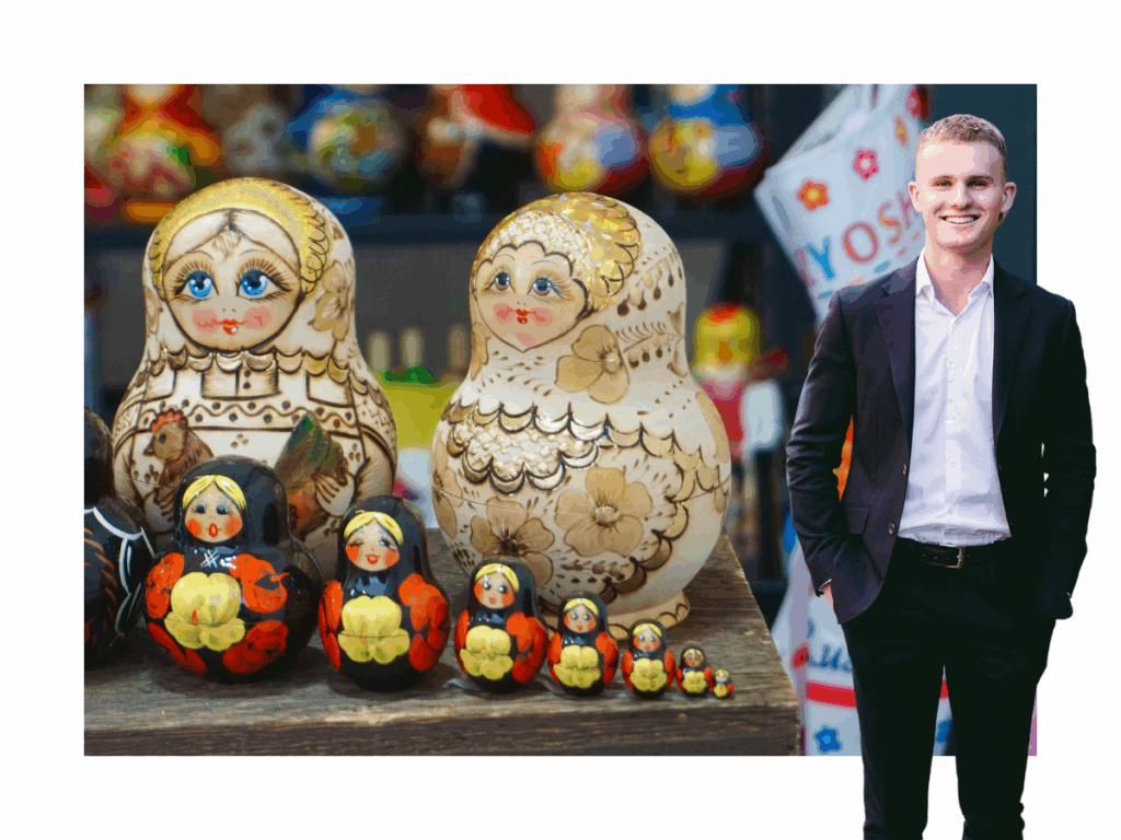 Pictured: Jayden Scot standing smiling in business attire, both with their hands in the pockets. Behind them is a close up image of Russian dolls.