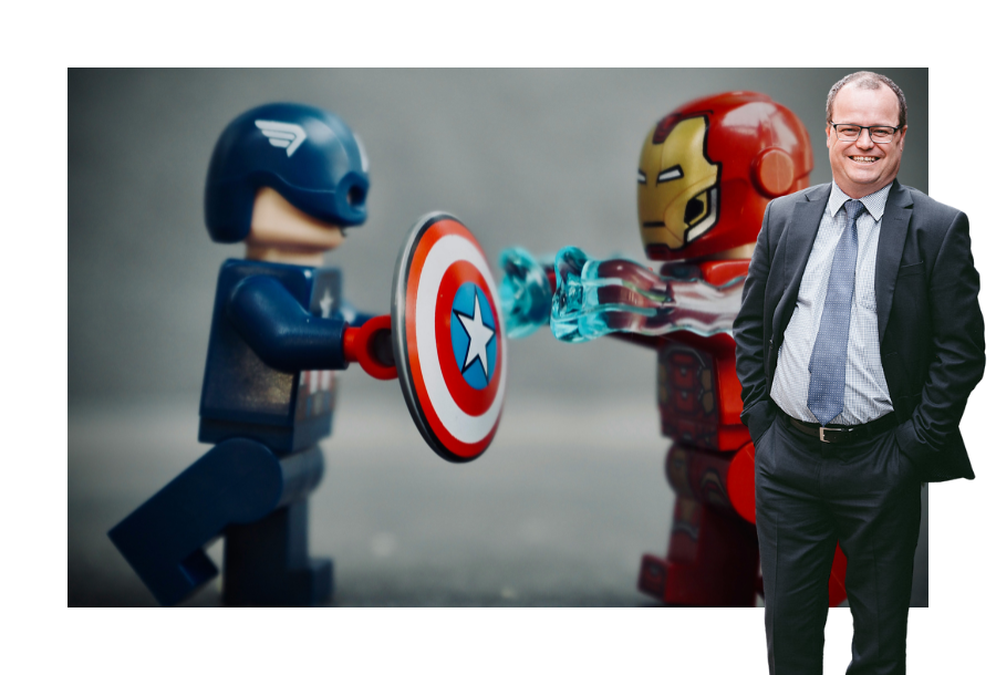 Pictured: Adam Wightman standing smiling wearing a black suit with a light blue shirt and blue tie with his hands in his trouser pockets. Behind him is an image of a lego Captain America and lego Iron man fighting.