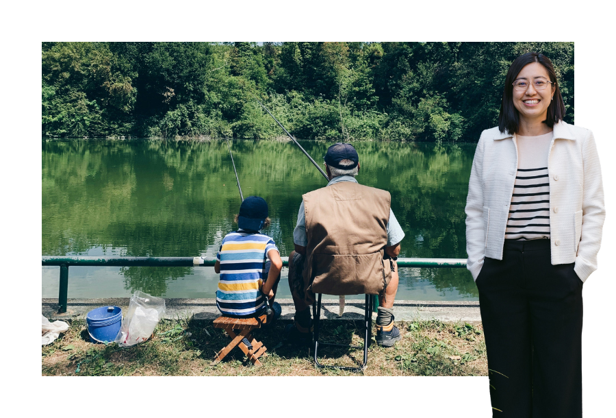Pictured: Helen Yau standing smiling with her hand in her pockets, wearing a beige cardigan and black trousers. Behind her is an image of a boy and his grandfather sitting fishing by a green lake, both are turned away from the camera facing the lake.