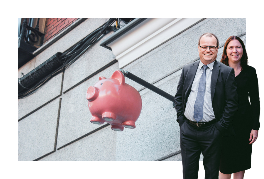 Pictured: Adam and Karlene Wightman standing smiling in business attire. Behind them is an image of a pink piggybank hanging from a pole coming off the external wall of a building.