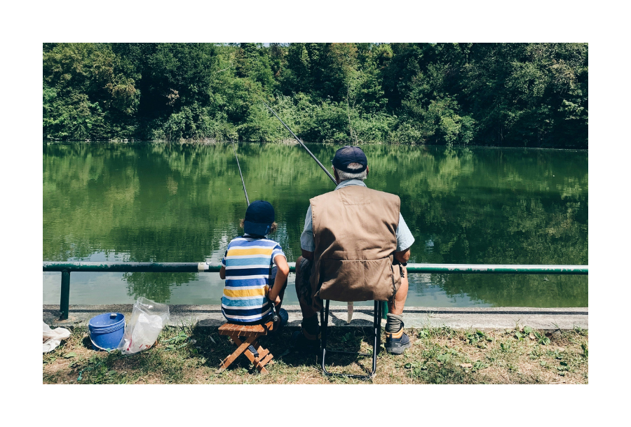 Pictured, a young pre-teen boy sitting with an older grandfather aged man. They are sitting with their backs in the sun, fishing out across a green lack.